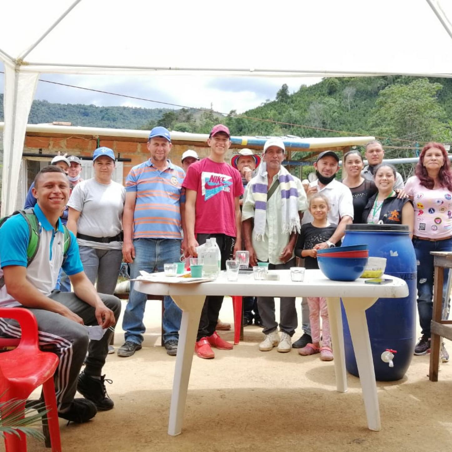 A group of coffee farmers stand around a white table with cups on it with green mountains in the background.