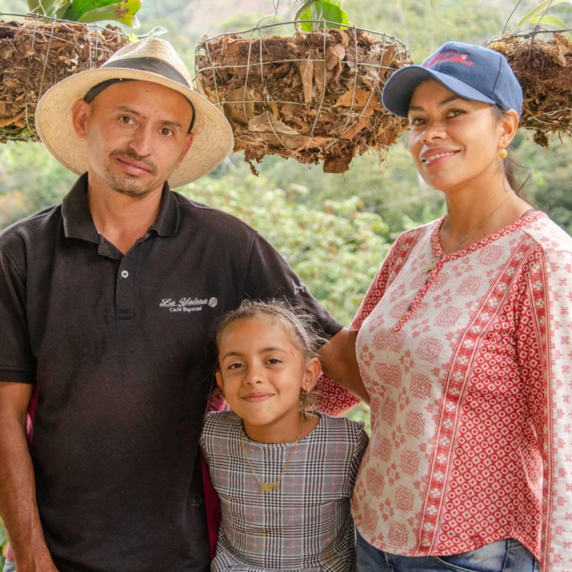 Elkin Diosa, a coffee farmer, stands for a portrait with his wife and daughter in front of hanging plants on their porch.